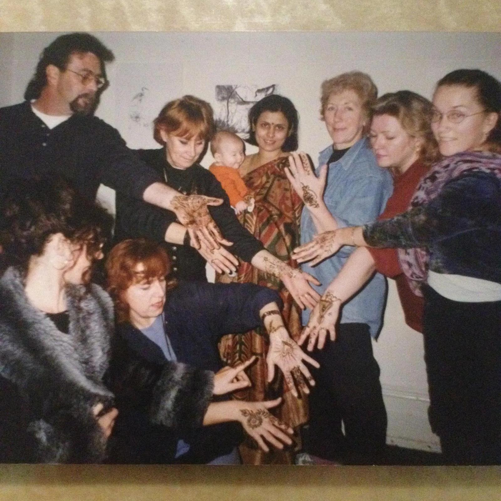 A 1990s henna workshop — eight adults with freshly hennaed hands raised toward the camera, a woman in a patterned sari at centre