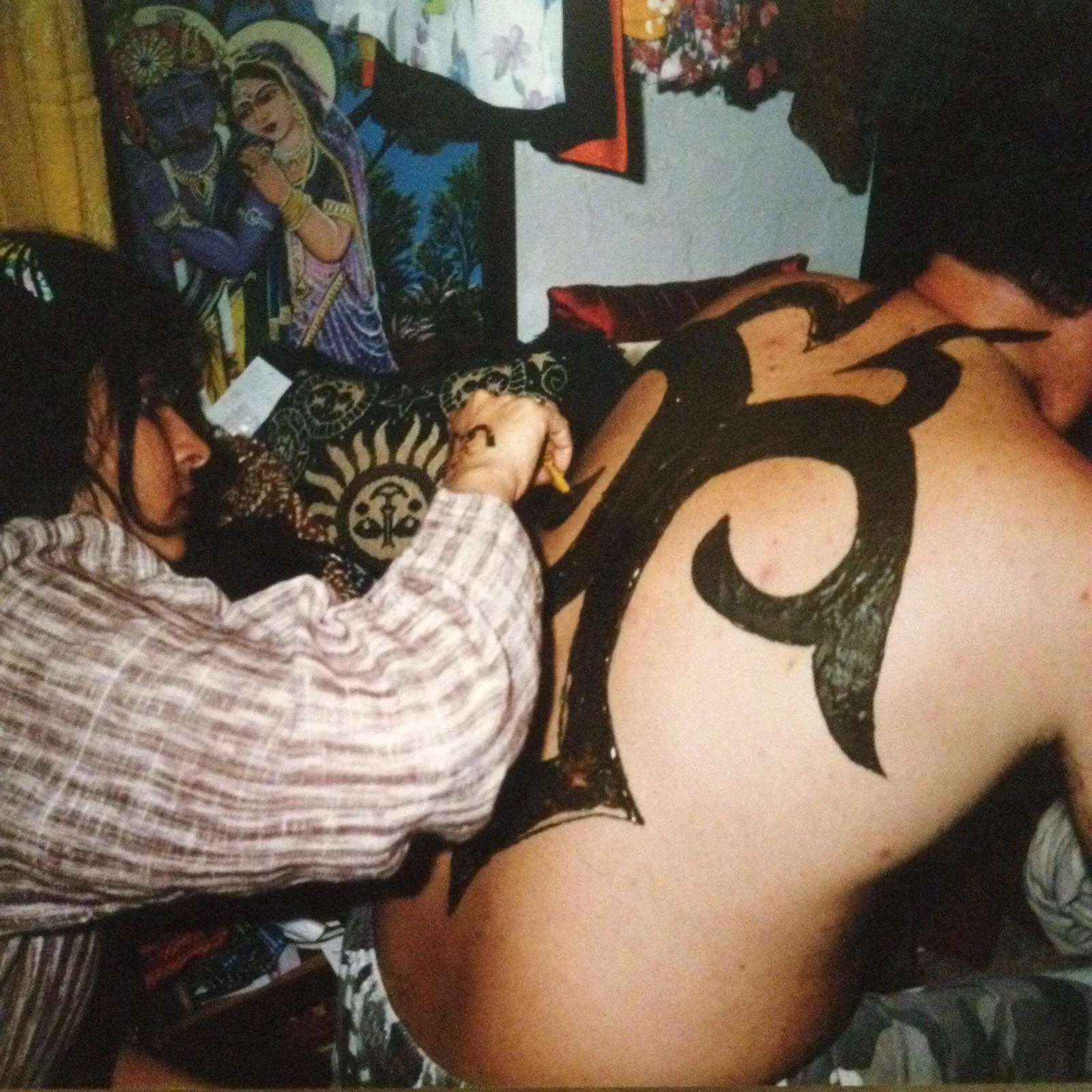 Vandana drawing a large black tribal henna design across a client's back, henna cone in hand, warm domestic lamplight